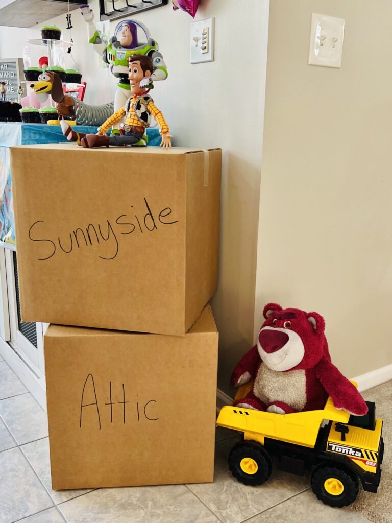 Toy Story's Lots-o'-Huggin Bear in a Tonka truck next to cardboard boxes labeled Sunnyside and Attic with Woody and Buzz toys on top