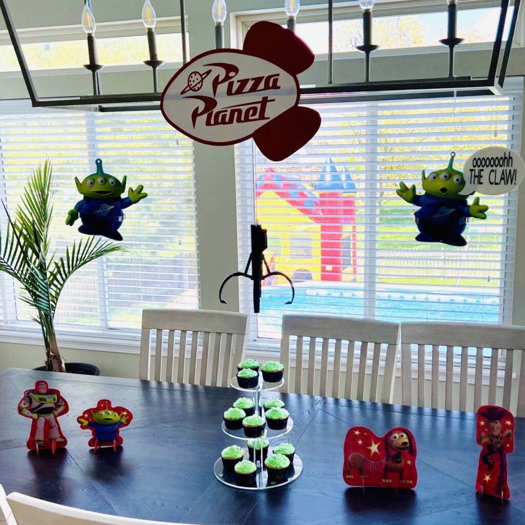 Toy Story birthday party table display with alien inspired cupcakes, pizza planet sign, claw, hanging honeycomb alien decorations and Toy Story cardboard table decorations
