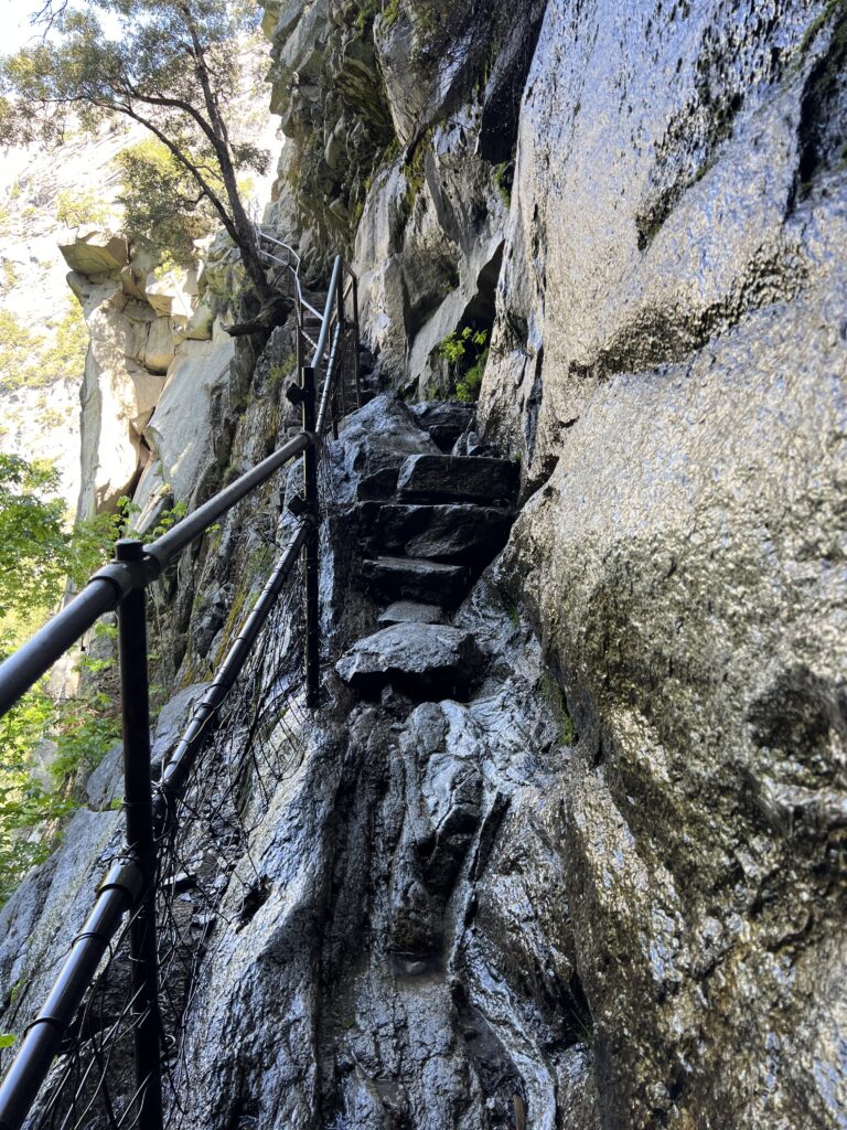 Steep narrow rock path with solid rock wall on one side and metal handrail along outer side of trail